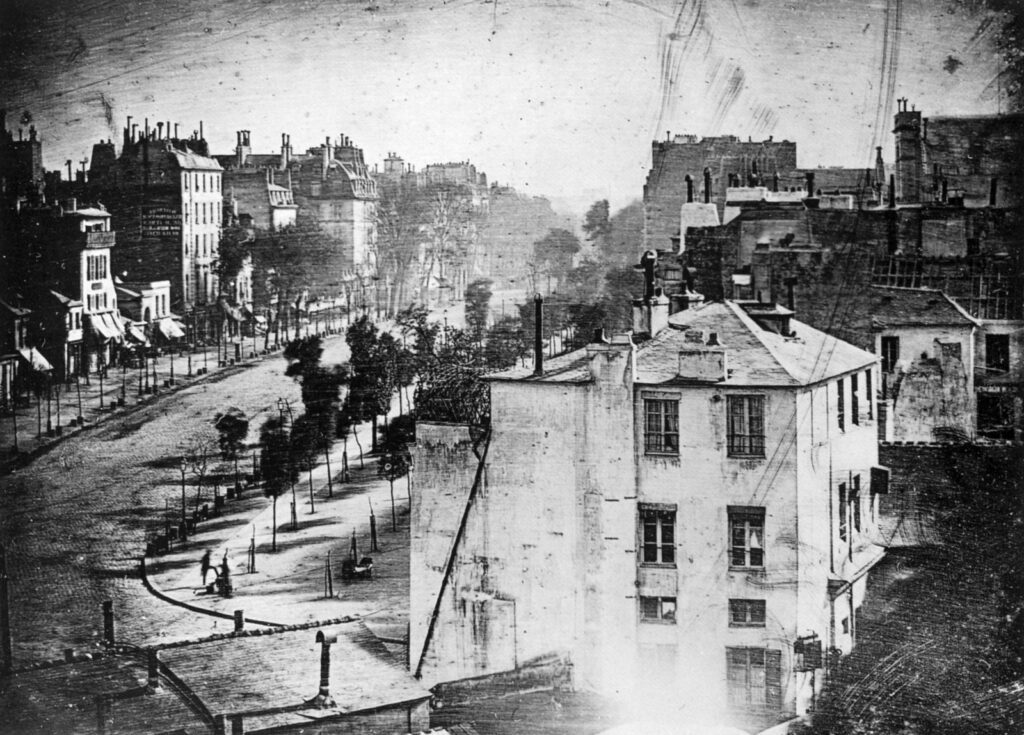 Daguerreotypie von Louis Daguerre einer Pariser Straßenansicht von 1838. Diese Aufnahme gilt als das älteste Foto, auf dem Menschen abgebildet sind, nämlich ein Schuhputzer und sein Kunde an der Straßenecke unten links. Quelle: aus "The Photography Book", Phaidon Press, London, 1997